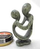 Front angle of serpentine stone sculpture showing the embracing figures and infinity shape next to a small can of shoe polish for size reference.