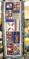 Full view of hand-woven textile with various traditional African geometric and symbolic motifs painted in blue, red, tan, black, and white, rectangular and measuring 60in x 20.5in.