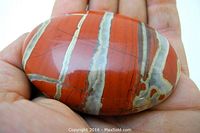 Close-up photo of the polished red jasper stone showing its orange-red base color with distinct white and gray stripes, resting on hand for scale.