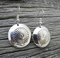 Close-up photo of a pair of round sterling silver Navajo earrings with detailed daisy flower design, shown hanging against weathered wood background.