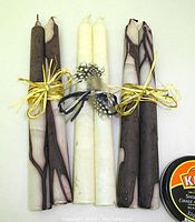 Photo showing three pairs of velvet dinner candles: two pairs are marbled dark brown and cream tones, and one pair is plain white. Each pair is tied with natural raffia and decorated with small feathers.