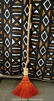 Front view of the full broom showing the long wooden handle and the fan-shaped orange-red brush corn bristles against a patterned black and white background.