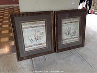 Photo of two framed prints angled on floor showing floral artwork and brown carved frames.