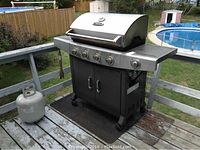 Full view of BBQTek propane grill with side burner, propane tank attached, utensils hanging from side rack, and protective mat underneath. Outdoor backyard setting with pool and grass visible.