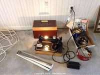 Singer sewing machine on wood base, with brown carrying case behind and collection of sewing accessories including tins of buttons and patterns visible to the side.