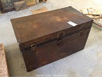 Exterior photo of a vintage metal trunk showing weathered surface, lock, clasps, and general wear.