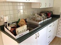 Wide view of kitchen countertop showing assorted mugs, glassware, vases in cardboard trays on a dark green counter with white cabinets below.