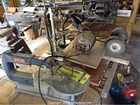 Ryobi scroll saw, homemade belt grinder laid on wooden surface with attached extra blades, visible electric cords.