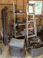 Photo showing scrap metal pile including metal shelving unit, aluminum ladder, assorted metal rods and parts, cast iron wood stove or component with rusted curved panel, and various pipes and scrap metal.