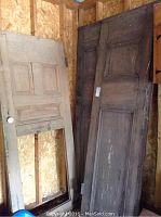 Three wooden doors leaning against wall, two large dark wood doors and one lighter wood door missing window pane.