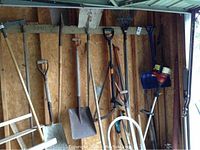 Various garden and hand tools hanging on wall, including shovels, rakes, sledge hammer, and gas weed trimmer.
