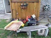 Wide view showing rusty metal wheelbarrow, black fertilizer spreader, various pots and gardening tools, and handcrafted alligator bench.