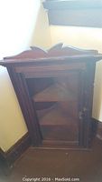 Full view of the corner display cabinet showing the dark wood finish, decorative top edge, glass front door, and interior shelves.