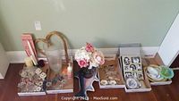 Wide view of the lot showing candle holders, glass vases, artificial flowers, multiple serving trays, picture frames, and eyeglasses all arranged on floor against wall.