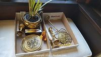 Brass planter with plant, two brass candle holders, decorative brass plate, and decorative brass stand arranged in boxes