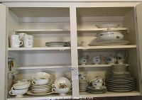 Full view of cabinet shelves holding stacked plates, cups, bowls, tureen and serving dishes, all white porcelain with floral designs and gold trim