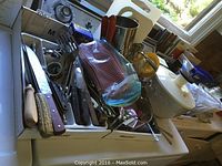 Collection of kitchen utensils on a stove including knives, forks, measuring cups, salad spinner, Pyrex bowls, and other kitchenware.