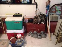 Wide view of sporting goods including coolers, dumbbells, hockey skates, walking sticks, gloves, and balls stacked against a wall.