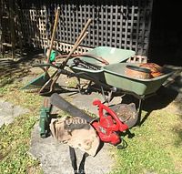 Wide angle photo showing two green metal wheelbarrows, Toro blower vac with collection bag, electric garden tools, rakes and sticks, some garden sprinklers and hardware, and an orange extension cord.