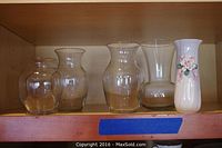 Five vases displayed on a wood shelf, four clear glass vases of different shapes and sizes, and one ceramic vase with floral decoration.