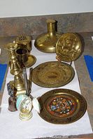 Photo of brass candlestick, small brass cup, brass bottle with narrow neck, decorative brass plates with maritime details, brass bell with figurine handle, trinket box.