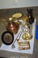 Overview of lot showing a small copper pitcher, heavy metal vase, plastic anniversary clock, brass candle snuffer, ornate letter opener, coffee spoon set in holder, small decorative box, and a small bell.