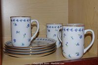 Four white ceramic mugs with blue wild flower pattern and gold rim, along with four matching cake plates stacked below them.