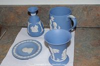 Photo of a set of blue Jasperware items including a small plate with Winston Churchill profile, a bud vase, and two vases/mugs with white relief decoration, placed on a countertop.