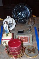 Wide view of all items in the lot showing decorative plate, candle, tea cup, glass, figurine, trinket box, butterfly box, tile and black base.