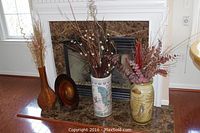 Wide photo of three decorative vases with dried floral arrangements placed on a marble fireplace hearth and wood floor background.