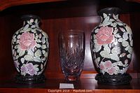 Two Sadek floral vases on dark wooden stands with one clear cut crystal vase in between, placed on wooden shelf