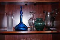Five glassware items on wooden shelf including tall frosted and clear swirl vase, blue glass decanter with stopper, ribbed clear glass vase, green textured vase, and etched clear glass pitcher.