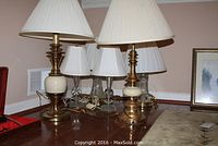 Photo showing six table lamps grouped with two tall brass and cream lamps and four smaller white shade lamps on wooden surface.