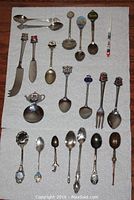 Full view of multiple collectible souvenir spoons laid out on a flat surface. Includes two spreading knives and forks among the spoons.