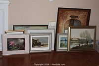 Overview showing six framed artworks propped against a wall on a wood floor, various sizes and frame finishes visible.