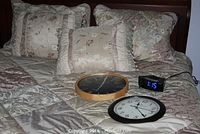 Queen size bed with floral patterned bedding including pillows, two wall clocks, and a digital alarm clock placed on bed for display.