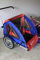 Full side-angle view of the InStep two-seater trailer showing blue and red fabric, clear plastic weather flaps, two wheels and rear jogging bar attached