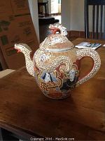 Porcelain teapot shown fully assembled on wooden table with intricate raised dragon and painted samurai figures.