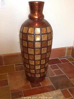 Front view of tall decorative plaster vase with grid square pattern in bronze and gold on tiled floor.