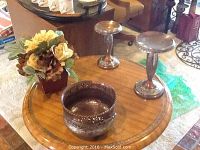 Tabletop setup showing two metal candle holders, faux flower arrangement, and small decorative metal bowl.