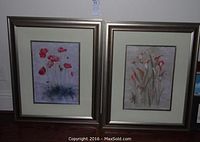 Two framed floral paintings showing poppies on the left and lilies on the right, both in matching metallic frames with cream mats and silver inner surrounds against a white wall.
