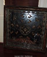 Square decorative mirror with ornate metallic frame and floral and scroll overlay design, placed on hardwood floor near door.