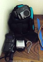 Photo showing Nikon Coolpix 990 digital camera inside a black and blue camera bag, with Gateway MPEG4 camcorder and Sony Video8 Handycam on wooden surface.