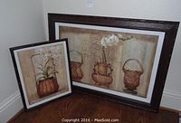Two coordinating framed wall art pieces arranged on floor showcasing white orchids and bamboo woven basket themes with dark brown frames.