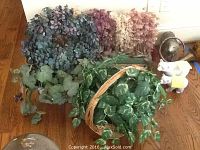 Photo shows eucalyptus wreath, dried flower bunches in pinks, faux green leafy plants in a basket and in pots on wood floor.