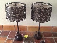 Pair of rattan style table lamps with dark woven shades and metal stands placed on a tiled surface against a wall.