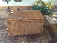 Photo showing rectangular wicker over wood trunk with woven texture, wood base, and metal handle. Trunk is on a rug with some decorative objects behind it.