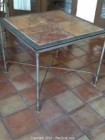 Square outdoor table with ceramic tile top, iron legs, and crossbar support. Earth tone tiles in browns and reds with visible wear and some damage.