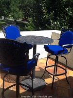 Wrought iron high-top round table with three matching high-top swivel chairs placed outdoors on tiled surface. Chairs have bright blue cushions on seat and back.