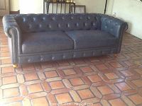 Front and side view of the Chesterfield tufted leather sofa with visible wear and cracks on the leather upholstery, showing rolled arms with nailhead trim and wooden legs with casters.
